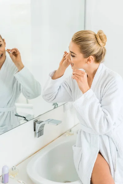 Selective focus of attractive and blonde woman in white bathrobe brushing teeth with dental floss — Stock Photo