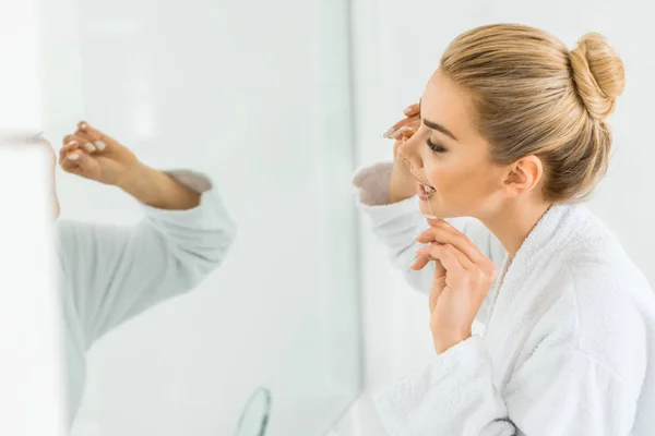 Foyer sélectif de femme attrayante et blonde en peignoir blanc brossant les dents avec fil dentaire — Photo de stock