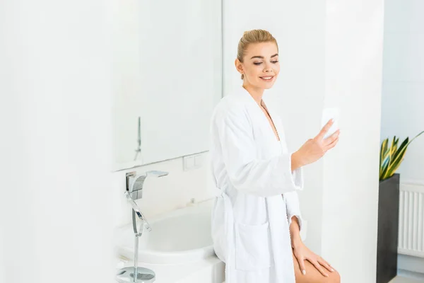 Привлекательная блондинка в белом халате делает селфи в ванной комнате — стоковое фото