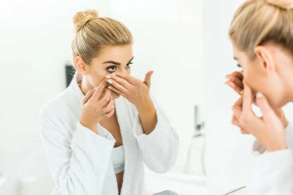 Messa a fuoco selettiva di attraente e bionda donna in accappatoio bianco fissaggio lente a contatto in bagno — Foto stock