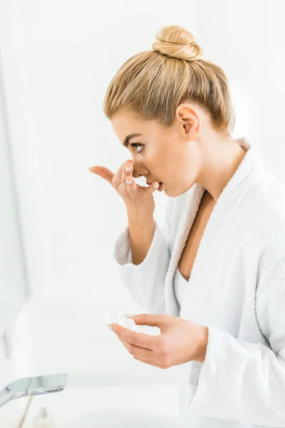 Mulher bonita e loira em roupão de banho branco anexando lente de contato no banheiro — Fotografia de Stock