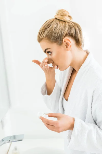 Bella e bionda donna in accappatoio bianco fissaggio lente a contatto in bagno — Foto stock