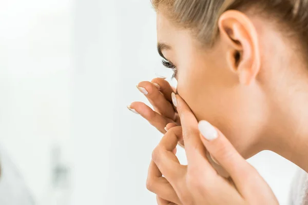 Selective focus of attractive and blonde woman in white bathrobe attaching contact lens — Stock Photo