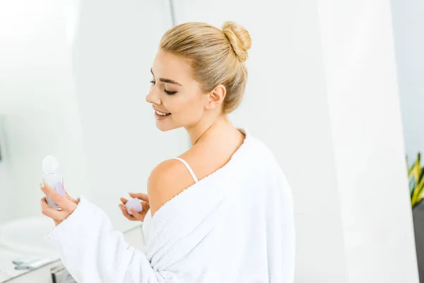 Femme blonde et souriante en peignoir blanc tenant déodorant dans la salle de bain — Photo de stock