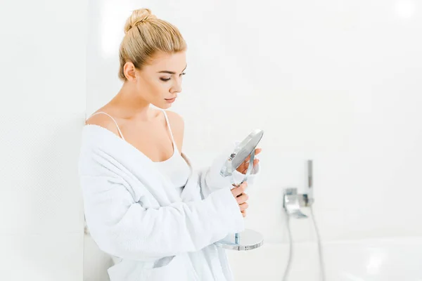 Attractive and blonde woman in white bathrobe looking at mirror in bathroom — Stock Photo