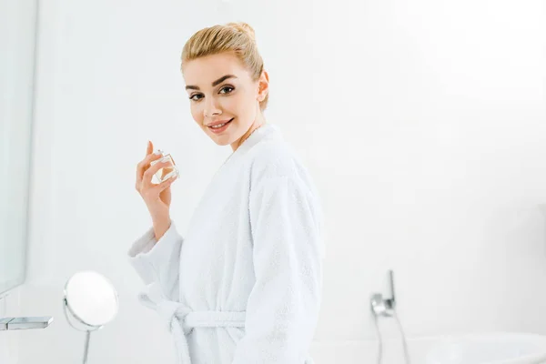 Beautiful and smiling woman in bathrobe holding perfume and looking at camera — Stock Photo