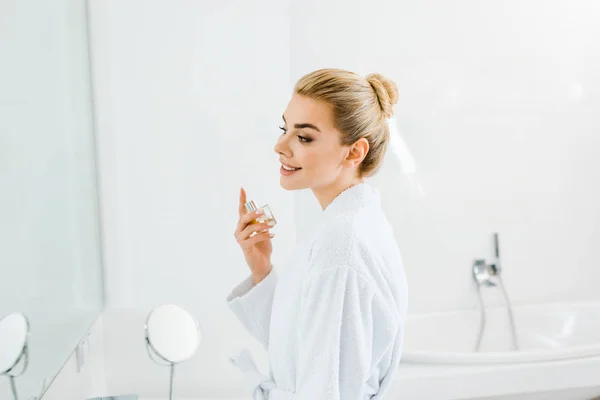 Schöne und lächelnde Frau im Bademantel mit Parfüm und Blick in den Spiegel — Stockfoto