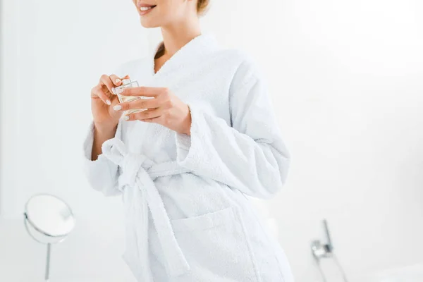 Cropped view of woman in white bathrobe holding perfume in bathroom — Stock Photo