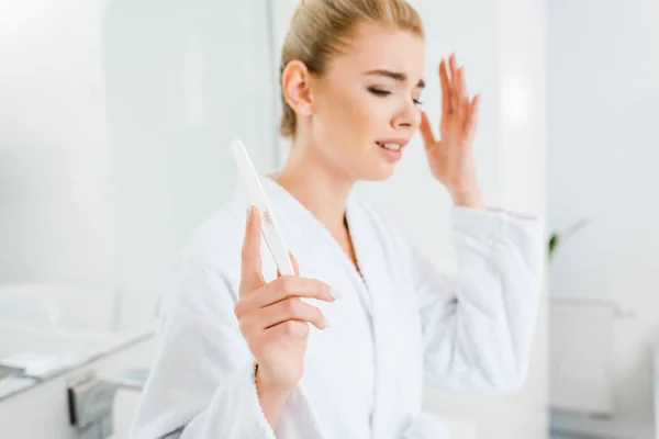 Селективный фокус грустной и блондинки в белом халате с пилкой для ногтей в ванной — стоковое фото