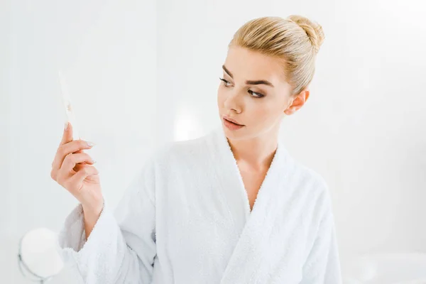 Mulher atraente e loira em roupão de banho branco segurando arquivo de unhas no banheiro — Fotografia de Stock