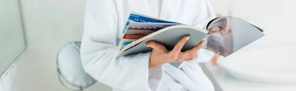 Panoramic shot of young adult woman in bathrobe holding magazine in bathroom — Stock Photo