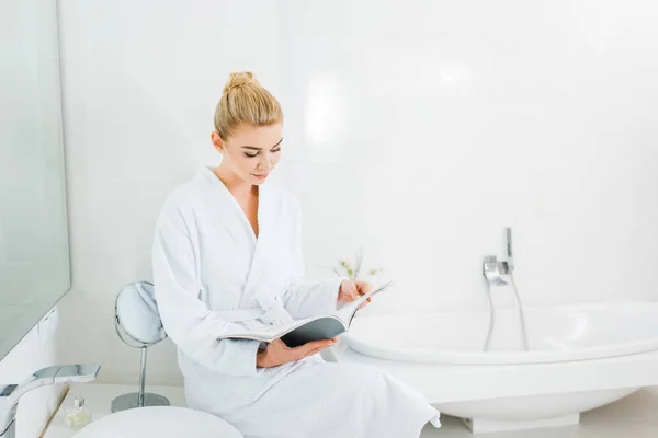 Attractive and blonde woman in white bathrobe reading magazine in bathroom — Stock Photo