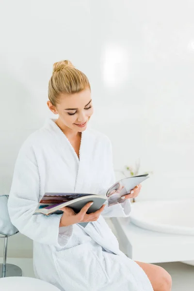 Attraente e sorridente donna in accappatoio bianco lettura rivista in bagno — Foto stock