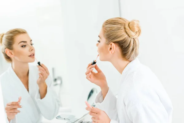 Selektiver Fokus der schönen und blonden Frau im weißen Bademantel, die Lipgloss im Badezimmer aufträgt — Stockfoto