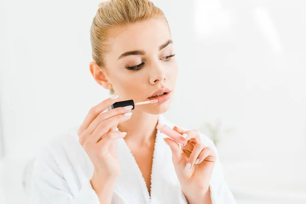 Mulher bonita e loira em roupão branco aplicando brilho labial no banheiro — Fotografia de Stock