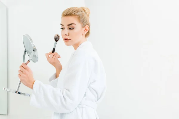 Beautiful and blonde woman in white bathrobe using cosmetic brush in bathroom — Stock Photo