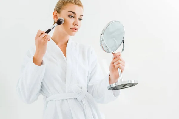Attractive and blonde woman in white bathrobe using cosmetic brush and holding mirror — Stock Photo
