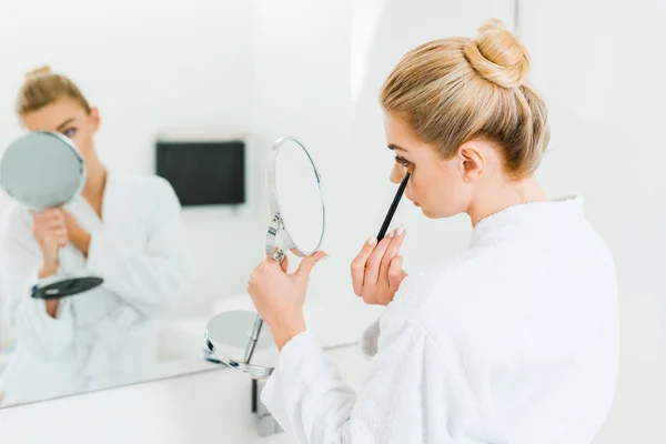 Selective focus of woman in white bathrobe applying eyeshadow with cosmetic brush — Stock Photo