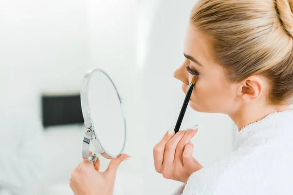 Selective focus of woman in white bathrobe applying eyeshadow with cosmetic brush in bathroom — Stock Photo