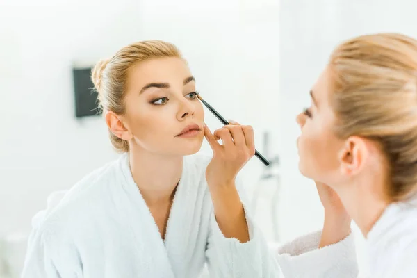 Selective focus of woman in white bathrobe applying eyeshadow with cosmetic brush — Stock Photo