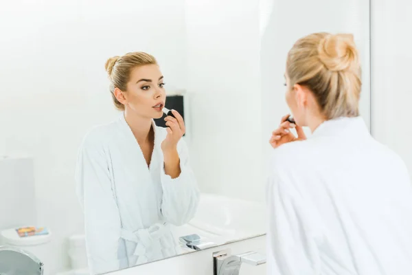 Selective focus of woman in white bathrobe applying lipstick and looking at mirror in bathroom — Stock Photo