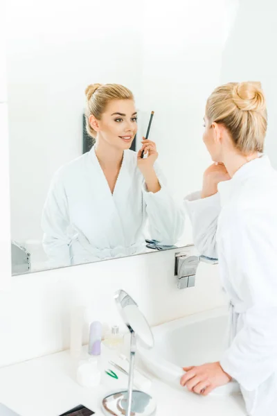 Selective focus of woman in white bathrobe holding cosmetic brush in bathroom — Stock Photo