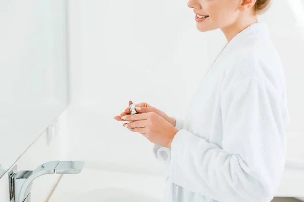 Vista recortada de la mujer en albornoz blanco celebración de lápiz labial en el baño - foto de stock