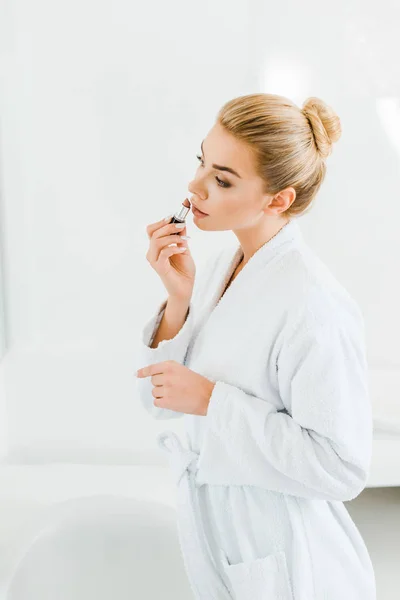 Hermosa y rubia mujer en albornoz blanco aplicando lápiz labial en el baño - foto de stock