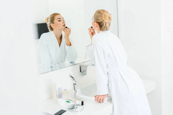 Enfoque selectivo de mujer hermosa y rubia en albornoz blanco aplicando lápiz labial en el baño - foto de stock