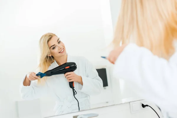 Foco seletivo da mulher bonita e loira em roupão de banho branco secando o cabelo no banheiro — Fotografia de Stock