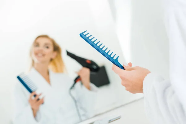 Selective focus of beautiful and blonde woman in white bathrobe holding hairdryer and comb — Stock Photo