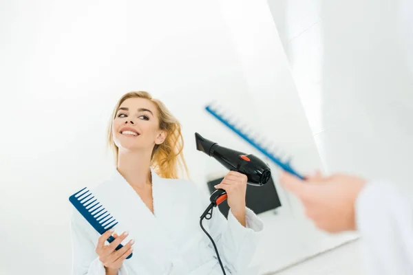 Selective focus of beautiful and smiling woman in white bathrobe holding hairdryer and comb — Stock Photo