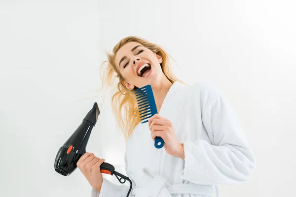 Beautiful and smiling woman in white bathrobe singing and holding hairdryer, comb — Stock Photo