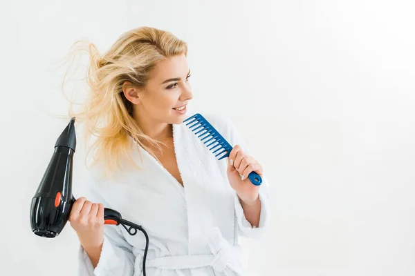 Beautiful and smiling woman in white bathrobe holding hairdryer and comb — Stock Photo