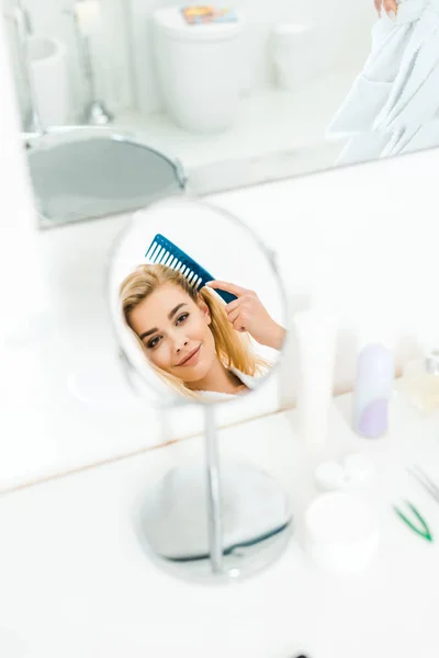 Selective focus of beautiful and smiling woman holding comb and looking at mirror — Stock Photo