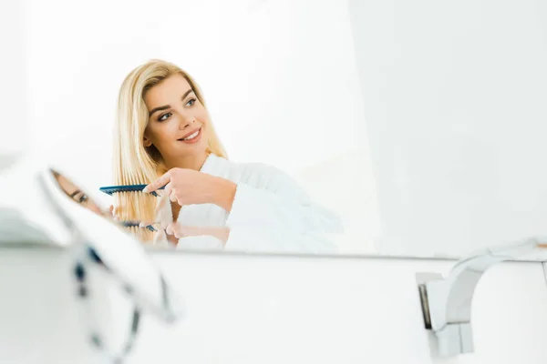 Schöne und lächelnde Frau im weißen Bademantel mit Kamm im Badezimmer — Stockfoto