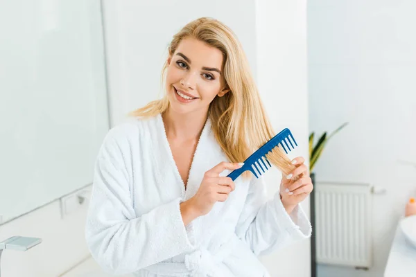 Schöne und lächelnde Frau im weißen Bademantel, die in die Kamera schaut und Kamm im Badezimmer benutzt — Stockfoto