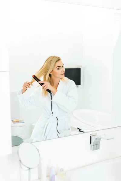 Mulher bonita e loira em roupão de banho branco usando ferro liso no banheiro — Fotografia de Stock