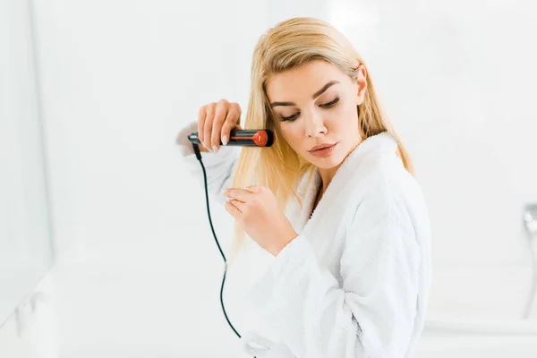 Belle femme blonde en peignoir blanc à l'aide de fer plat et regardant vers le bas dans la salle de bain — Photo de stock