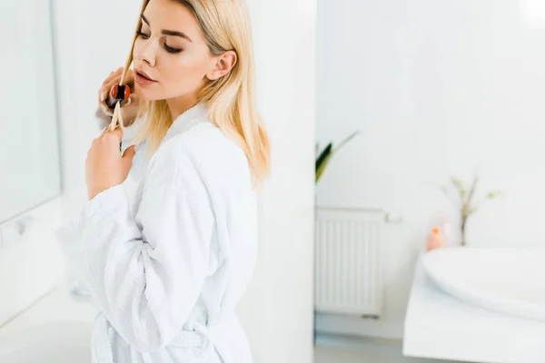 Beautiful and blonde woman in white bathrobe using flat iron and looking away in bathroom — Stock Photo