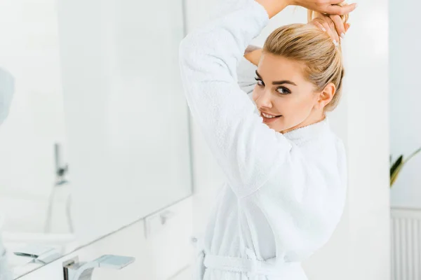 Bella donna in accappatoio bianco giocare con i capelli e guardando la fotocamera in bagno — Foto stock