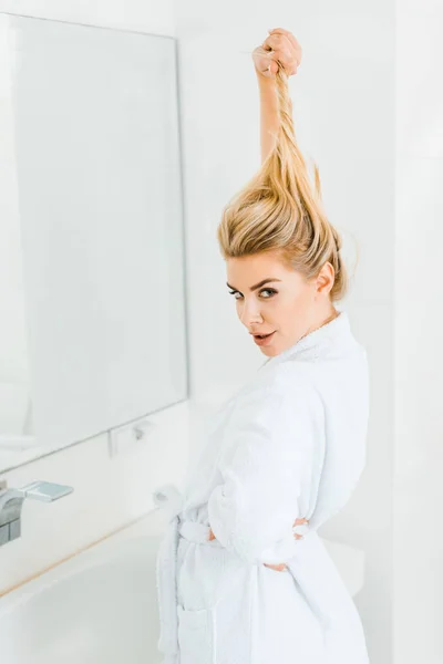 Attractive woman in white bathrobe playing with hair and looking at camera in bathroom — Stock Photo