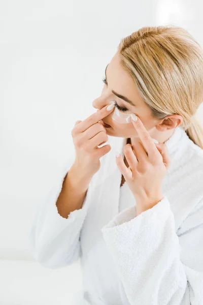 Mujer rubia y atractiva en albornoz blanco aplicando parches para los ojos en el baño - foto de stock