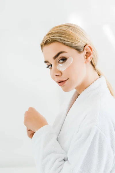 Beautiful and blonde woman in white bathrobe with eye patches on face looking at camera in bathroom — Stock Photo