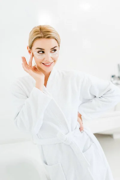 Beautiful and smiling woman in white bathrobe with eye patches on face looking away in bathroom — Stock Photo