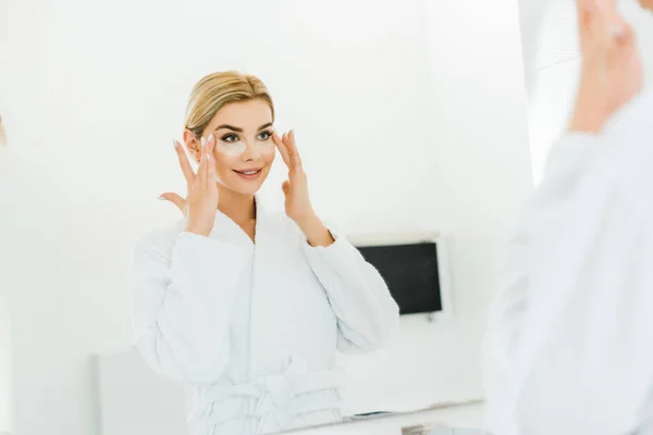 Selective focus of beautiful and blonde woman in white bathrobe applying eye patches in bathroom — Stock Photo