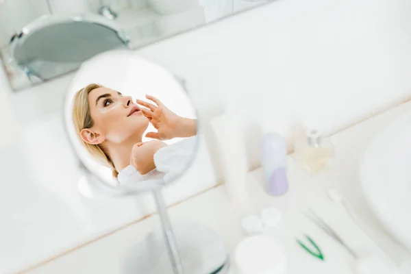 Selektiver Fokus der attraktiven und blonden Frau im weißen Bademantel, die Augenklappen im Badezimmer aufträgt — Stockfoto