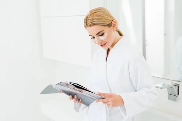 Beautiful and smiling woman in white bathrobe with eye patches on face reading magazine — Stock Photo