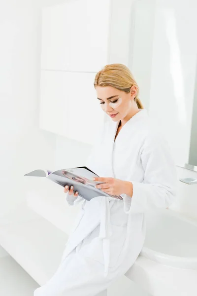 Beautiful and blonde woman in white bathrobe with eye patches on face reading magazine — Stock Photo