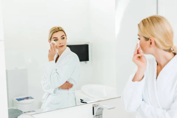 Selective focus of beautiful and blonde woman in white bathrobe using cotton pad and looking at mirror — Stock Photo